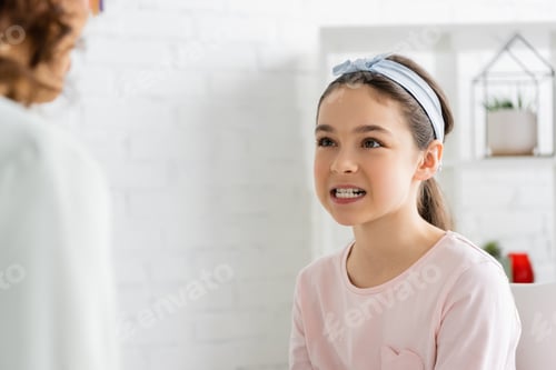 Preview: Preteen girl showing teeth near blurred speech therapist in consulting room