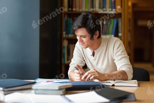 Preview: Male student leaning and writing in notebook with pen at library table with textbooks, copy space