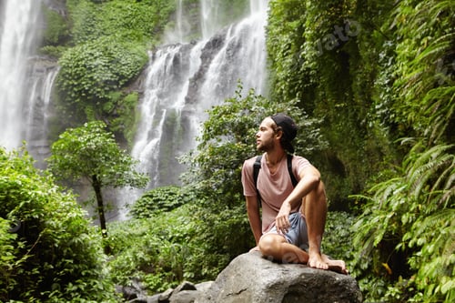 Preview: Young Caucasian barefooted male tourist with backpack sitting on rock surrounded by rainforest and a