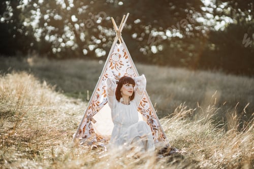 Preview: Native American stunning woman, sitting near stylish boho tee pee wigwam tent in summer field