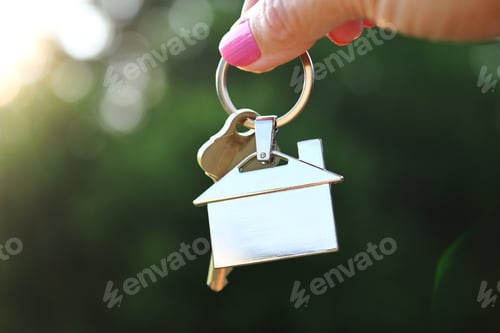 Preview: Female hand of realtor holding a set of house keys with home keychain outside during golden hour.