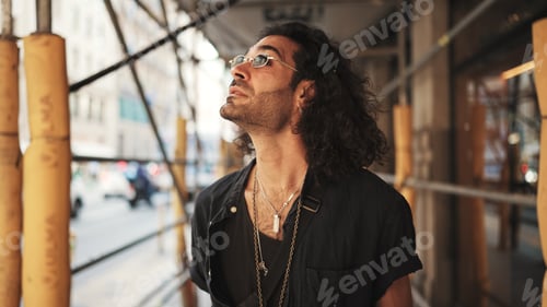 Preview: Young italian guy with long curly hair and stubble walks along the fenced sidewalk and looks around.