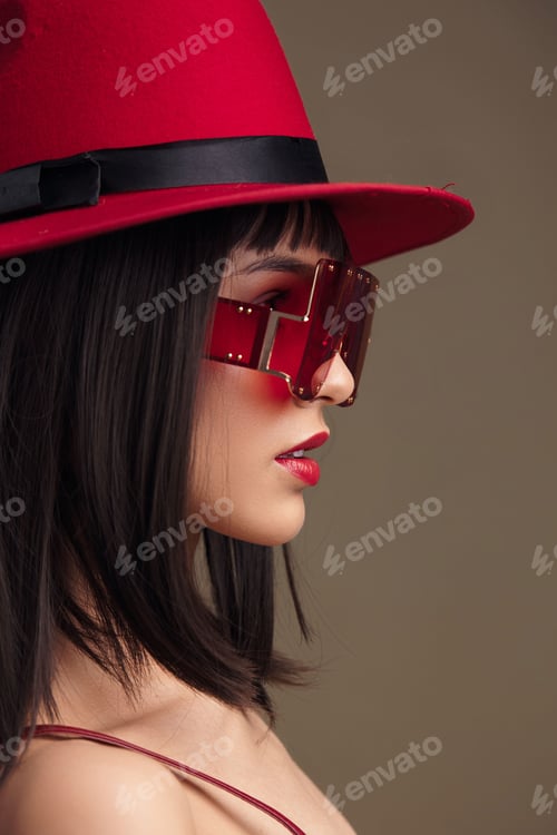 Preview: Woman in red dress and fedora, wearing sunglasses on her head, standing confidently against a bright