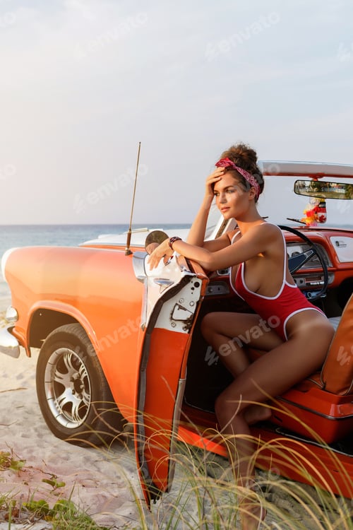 Preview: Sexy woman wearing red swimsuit in retro cabriolet car on the beach.