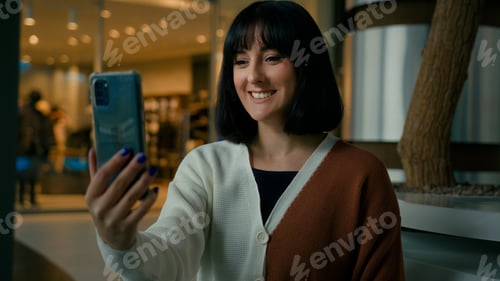 Preview: Smiling positive talking to smartphone webcam woman has video call indoors in shopping mall waving