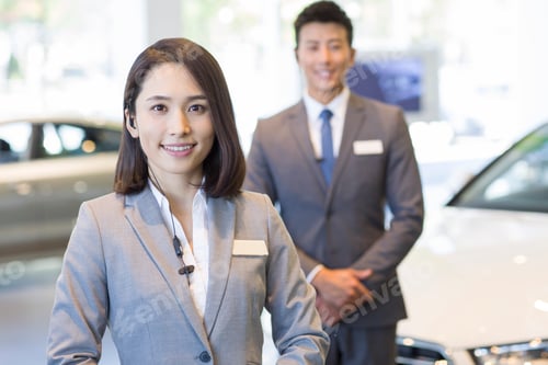 Preview: Confident salespeople standing with new cars in showroom