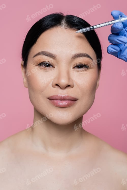 Preview: Asian woman with naked shoulders looking at camera near cosmetologist holding syringe isolated on