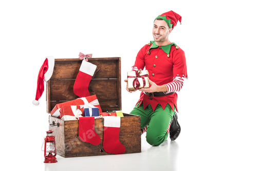 Preview: smiling man in christmas elf costume sitting near wooden chest full of presents and holding gift