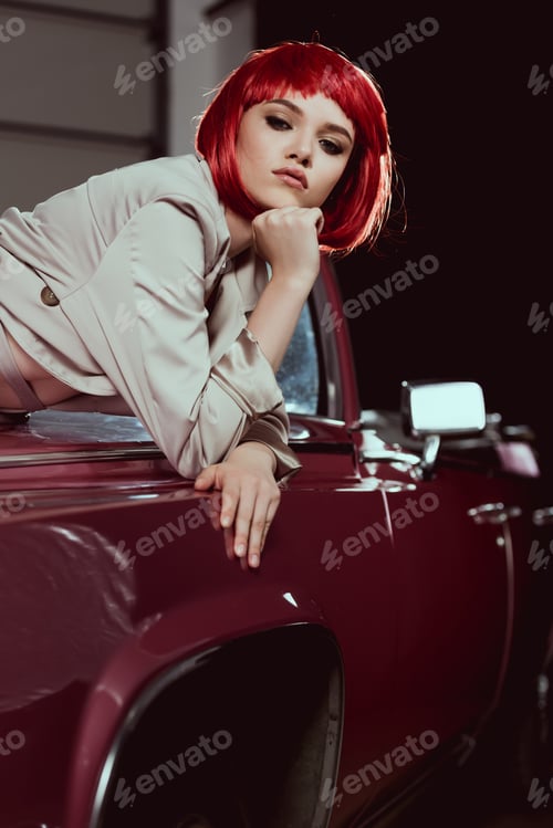 Preview: attractive young woman in red wig and trench coat looking at camera while leaning at classic car
