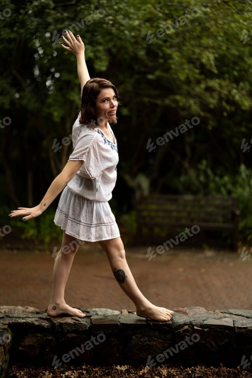 Preview: Female standing on a stone fence in a scenic outdoor setting, surrounded by lush greenery and trees