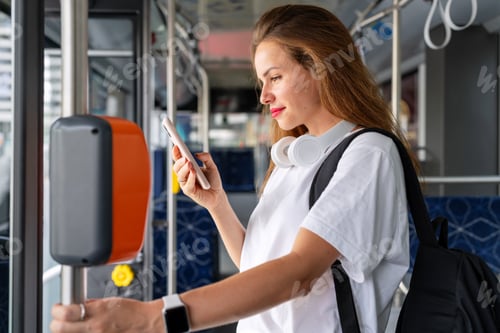 Preview: Brunette woman in white tee shirt standing inside bus and using her smart phone.