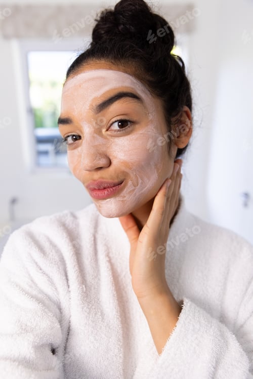 Preview: Portrait of biracial woman in bathrobe with face mask in sunny bathroom