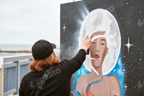 Preview: Female artist in black cap is painting picture with paint spray can spraying it onto canvas