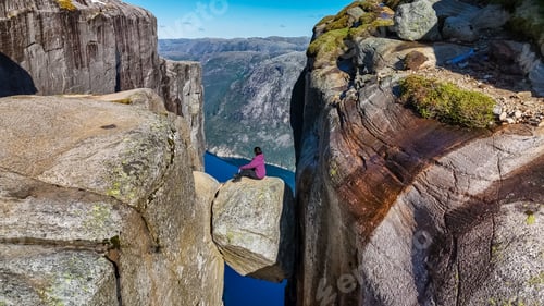 Preview: A lone hiker sits on the edge of Kjeragbolten, Norway, a famous cliff in Norway, overlooking a