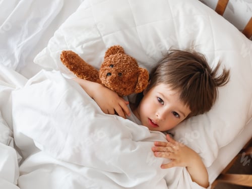 Preview: Toddler lies in bed with cute teddy bear. Little boy under white blanket with fluffy toy.