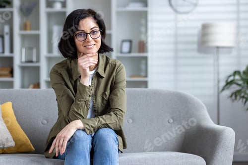 Preview: Portrait of a young smiling woman sitting on the couch at home, leaning her head on her hand and