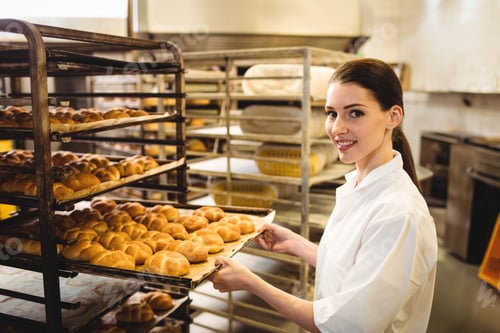 Preview: Female baker removing tray of golden brown rolls from metal rack in commercial bakery, copy space
