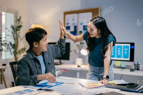 Preview: Two coworkers are giving each other a high five over a table with paperwork and digital devices, cel
