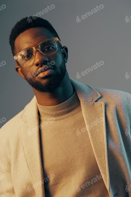 Preview: Stylish young Black man with glasses in a cozy beige sweater and light gray coat, expressing