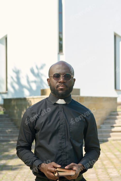 Preview: Young serious priest in black shirt with clerical collar holding Holy Bible