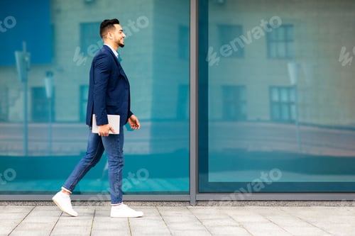 Preview: Cheerful arab businessman with pad walking by street, full length
