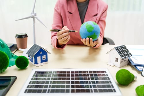 Preview: A businesswoman in a pink suit stands at a white desk with a world model, electric car model