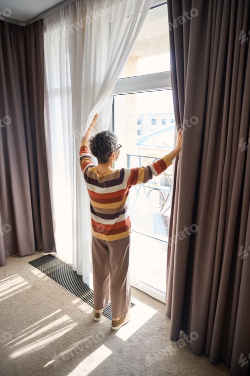 Preview: Senior woman opening curtains and enjoying sunlight standing in hotel room