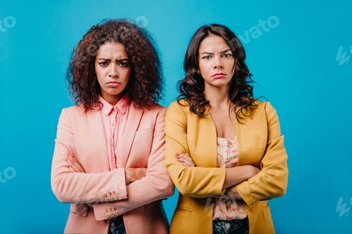 Preview: Disappointed girls looking to camera. Indoor photo of two brunette women standing on studio backgro