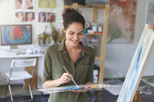 Preview: Portrait of cute female artist working in workshop, painting with brush and watercolors, standing ne