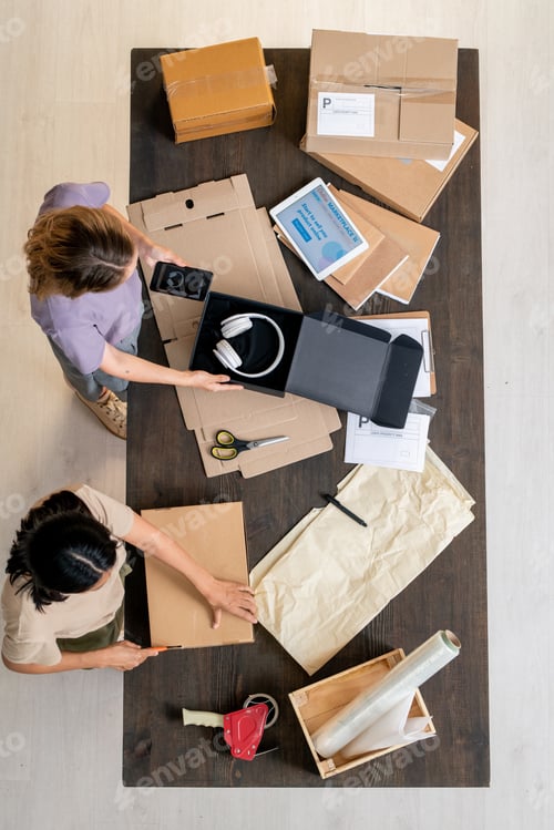 Preview: Top view of two young women standing by table and packing orders of clients