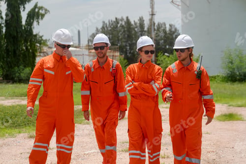 Preview: Workers in orange coveralls discuss operations near industrial facility on sunny day