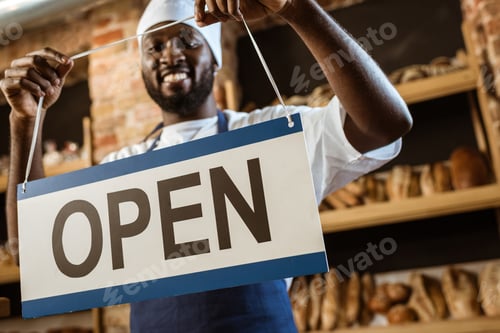 Preview: happy african american baker holding open sign at pastry store