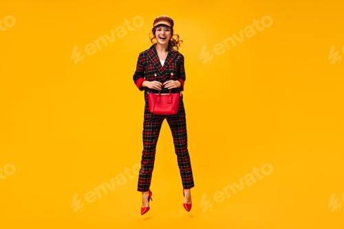 Preview: Cheerful lady in stylish outfit holds red handbag. Modern happy young woman with curly hair in cap,