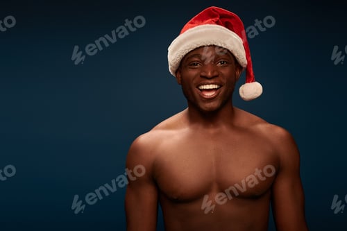 Preview: happy and shirtless african american guy in santa hat smiling at camera on navy blue backdrop