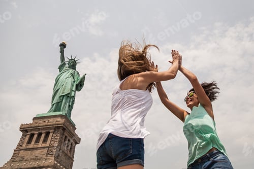 Preview: Two Friends Giving a High Five Jumping in the Air besides the Statue of Liberty
