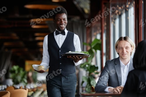 Preview: Smiling waiter bringing food on tray to guests enjoying dinner