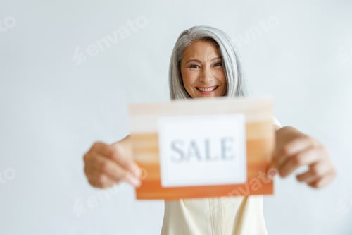 Preview: Happy mature Asian womanshows Sale sign standing on light grey background