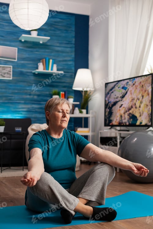 Preview: Peaceful senior woman with closed eyes sitting on yoga mat meditating during wellness workout