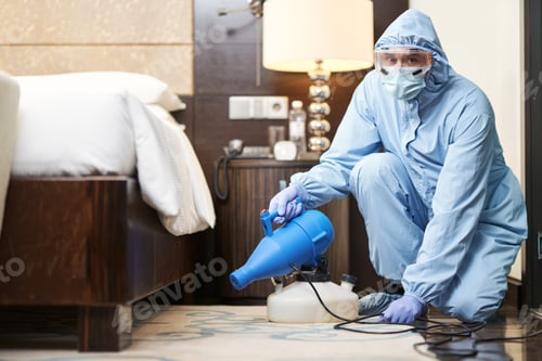 Preview: Worker in biohazard suit and mask disinfecting bedroom