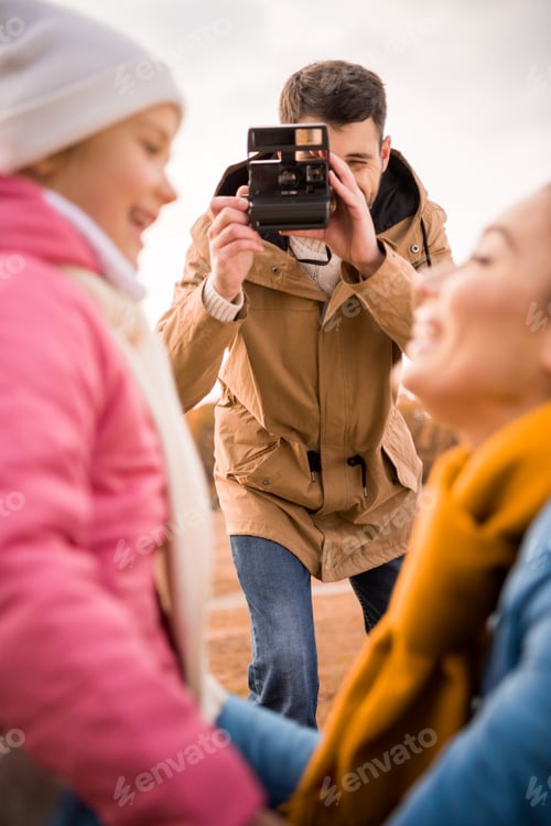 Preview: Young man photographing happy mother and daughter with instant camera outdoors
