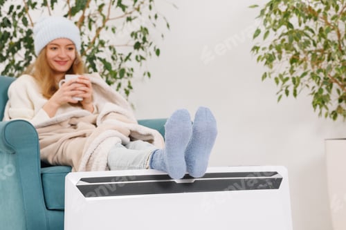 Preview: Young woman with warm plaid and tea cup sitting near electric convector heater in bedroom