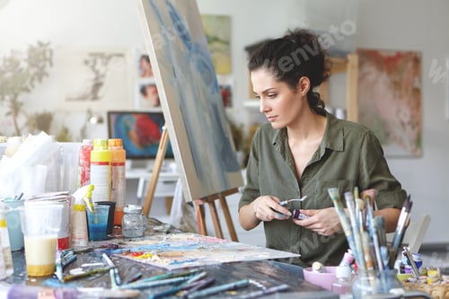 Preview: Young female painter sitting at table surrounded with various paint brushes and watercolors while cr