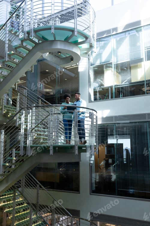 Preview: African american professionals standing on staircase discussing together over laptop at workplace