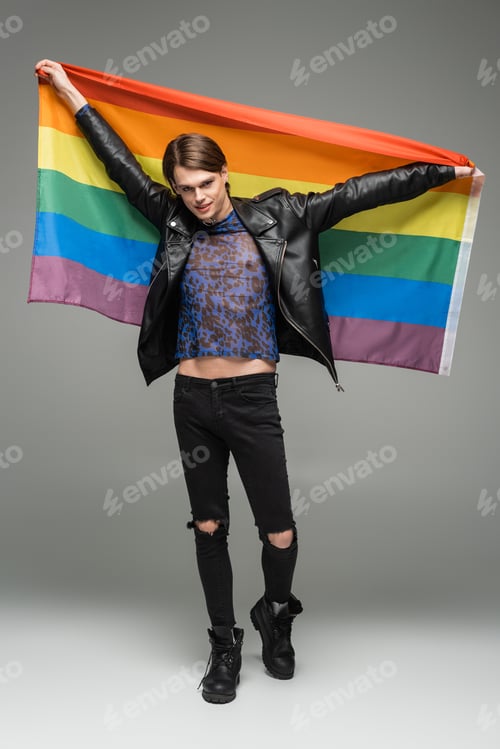 Preview: full length of smiling pangender person in black ripped pants and leather jacket holding rainbow