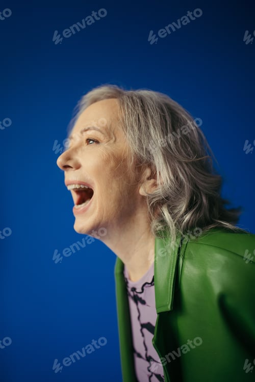 Preview: astonished senior woman in green leather jacket looking away isolated on blue