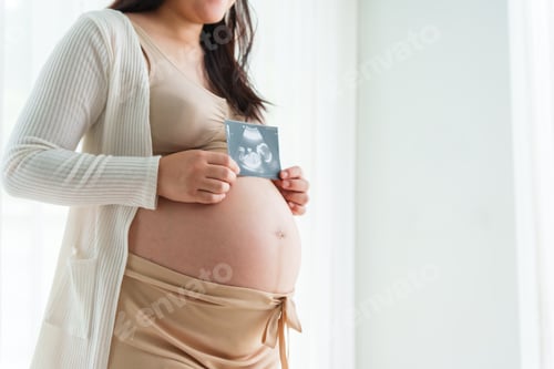 Preview: An Asian young pregnant wife relaxes at home, hand on belly, filled with love and anticipation.