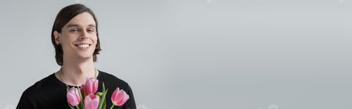 Preview: Smiling brunette man holding tulips and looking at camera isolated on grey, banner