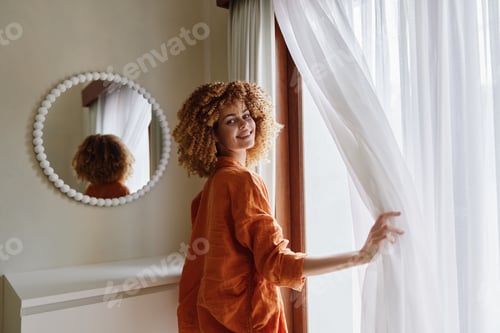 Preview: Beautiful young woman with curly hair wearing an orange outfit, smiling gently while standing near a