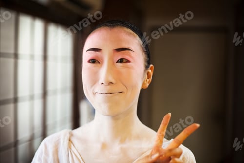 Preview: Geisha woman with traditional white face makeup and heavy eyeliner holding up two fingers in a