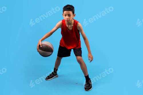 Preview: Boy in red uniform training, playing basketball, dribbling over blue studio background.